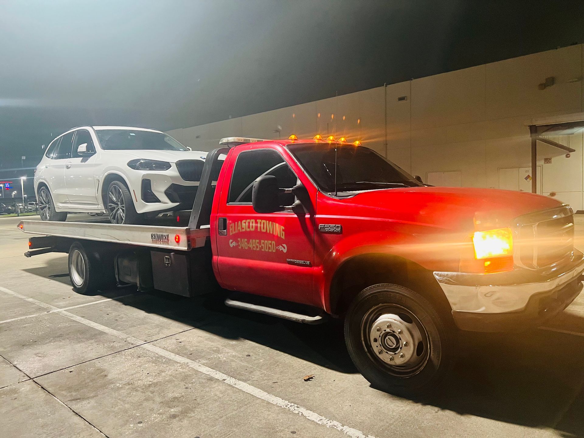 Red tow truck carrying a white SUV on a flatbed at night with headlights on