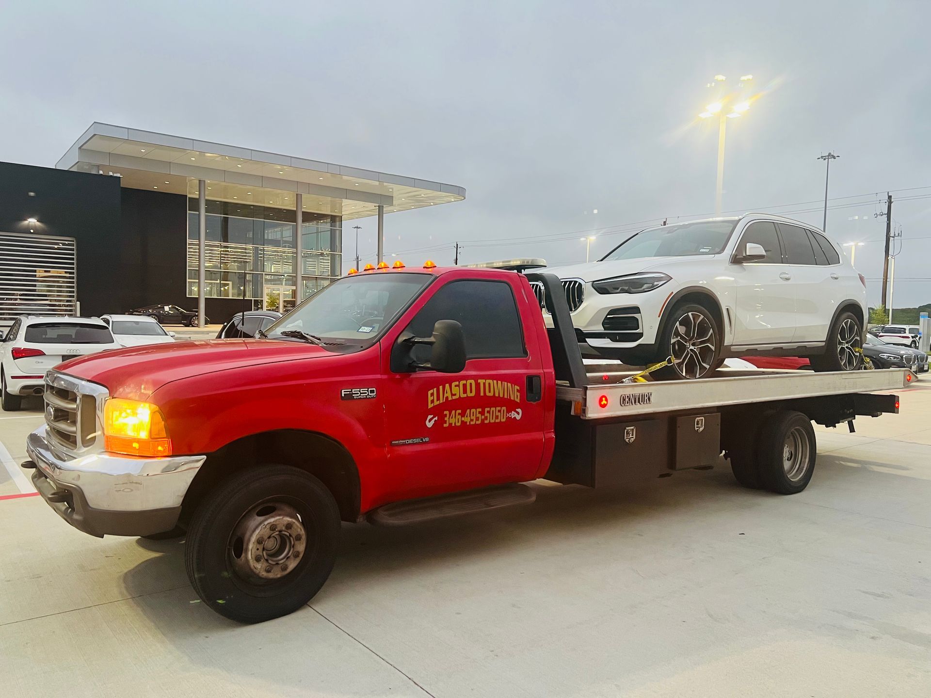 Red tow truck carrying a white SUV in a parking lot at dusk