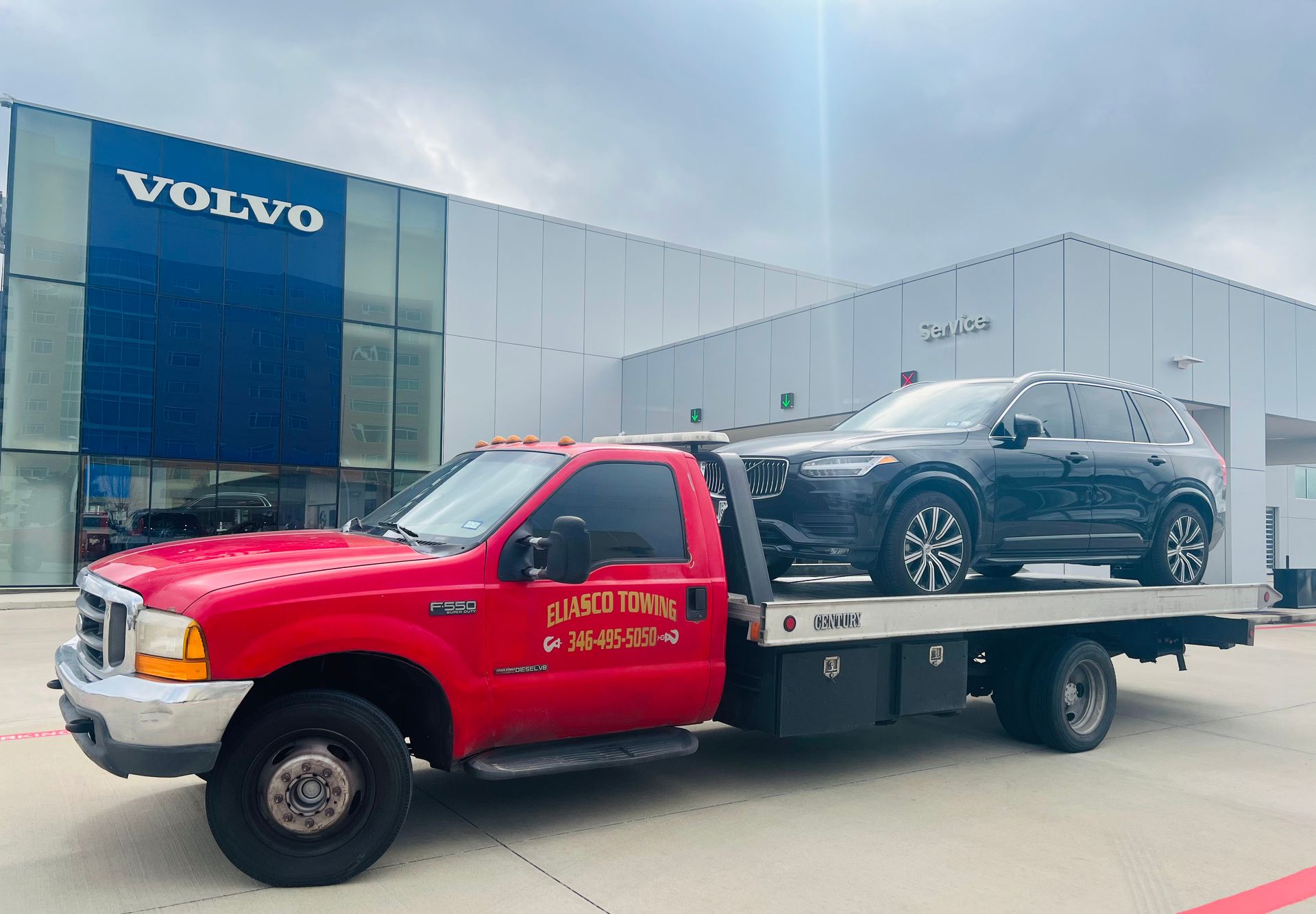 Red tow truck hauling a dark SUV outside a Volvo dealership