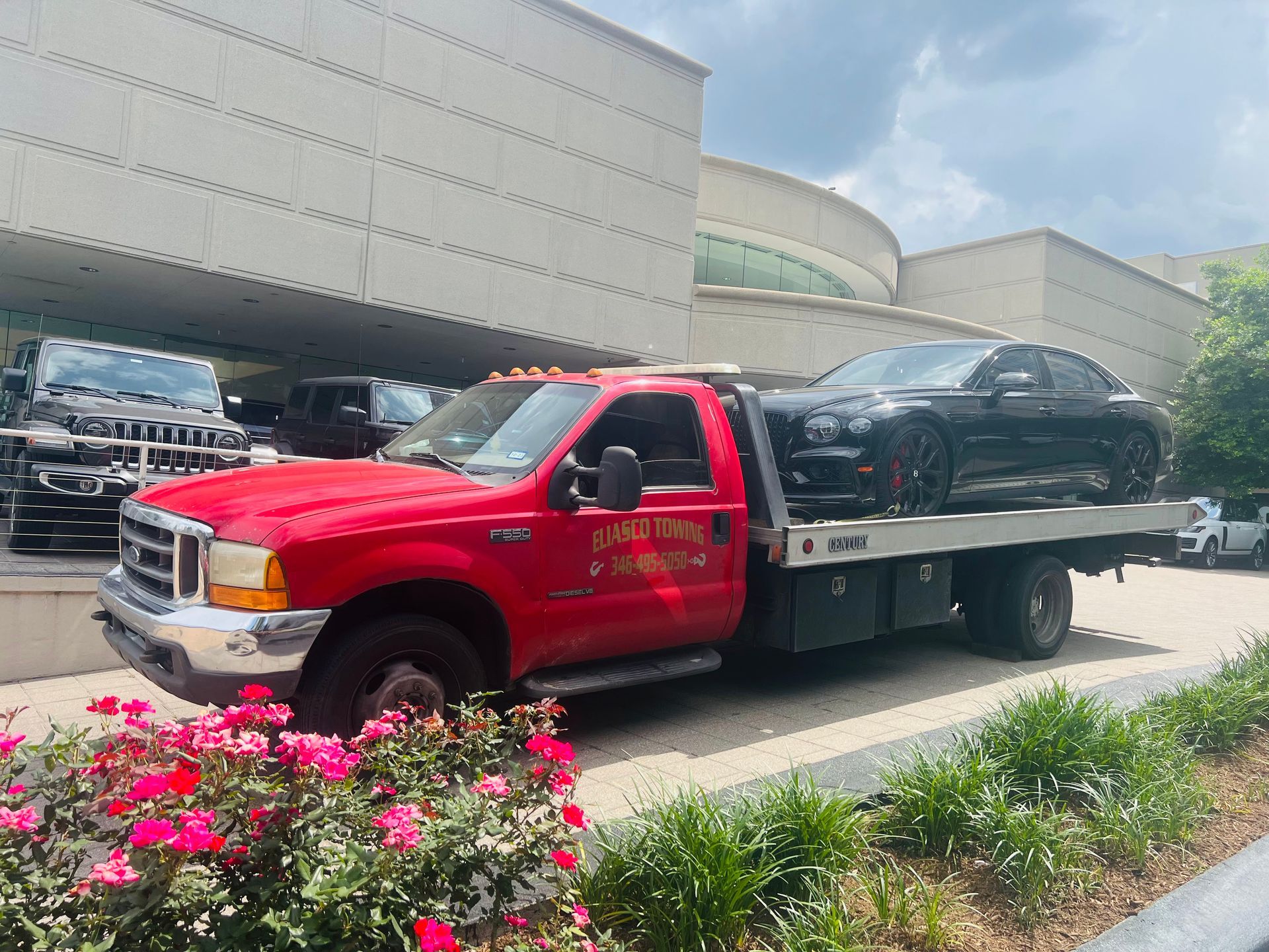 Red flatbed tow truck carrying a black car, parked outside a building with flowers in front.
