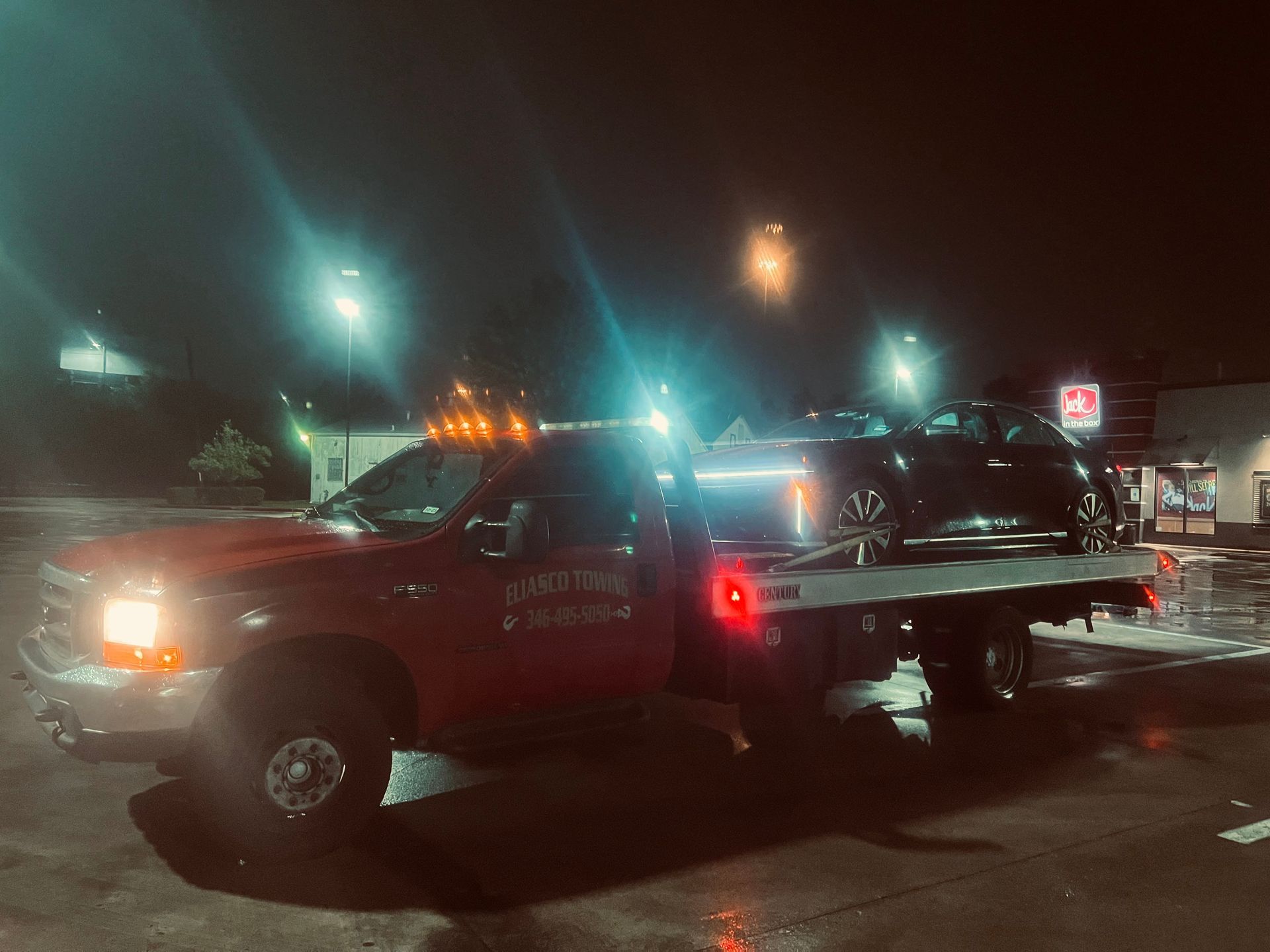 Tow truck hauling a black car at night in a parking lot, with bright streetlights and store signs.