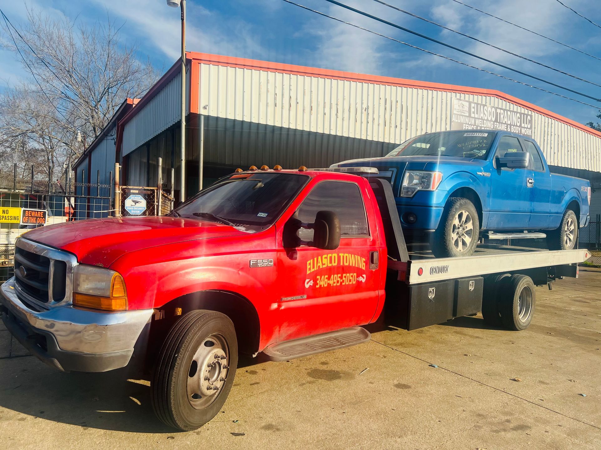 Red tow truck carrying a blue pickup on a flatbed outside a building