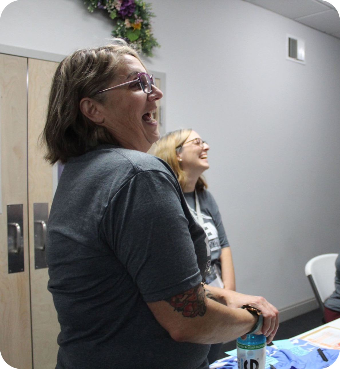 Two women laughing at a table in a gray-walled room, one holding a water bottle.