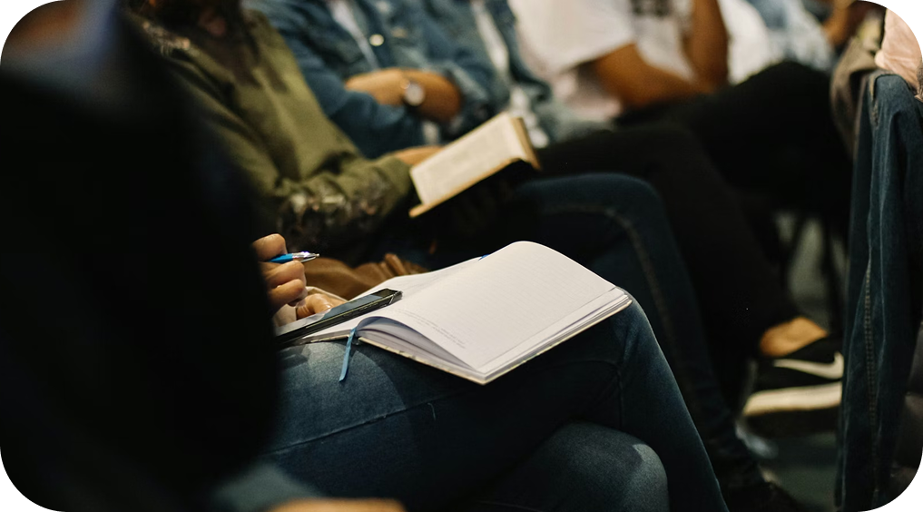 Hands holding open notebooks in a dim audience seating area, suggesting a lecture or workshop