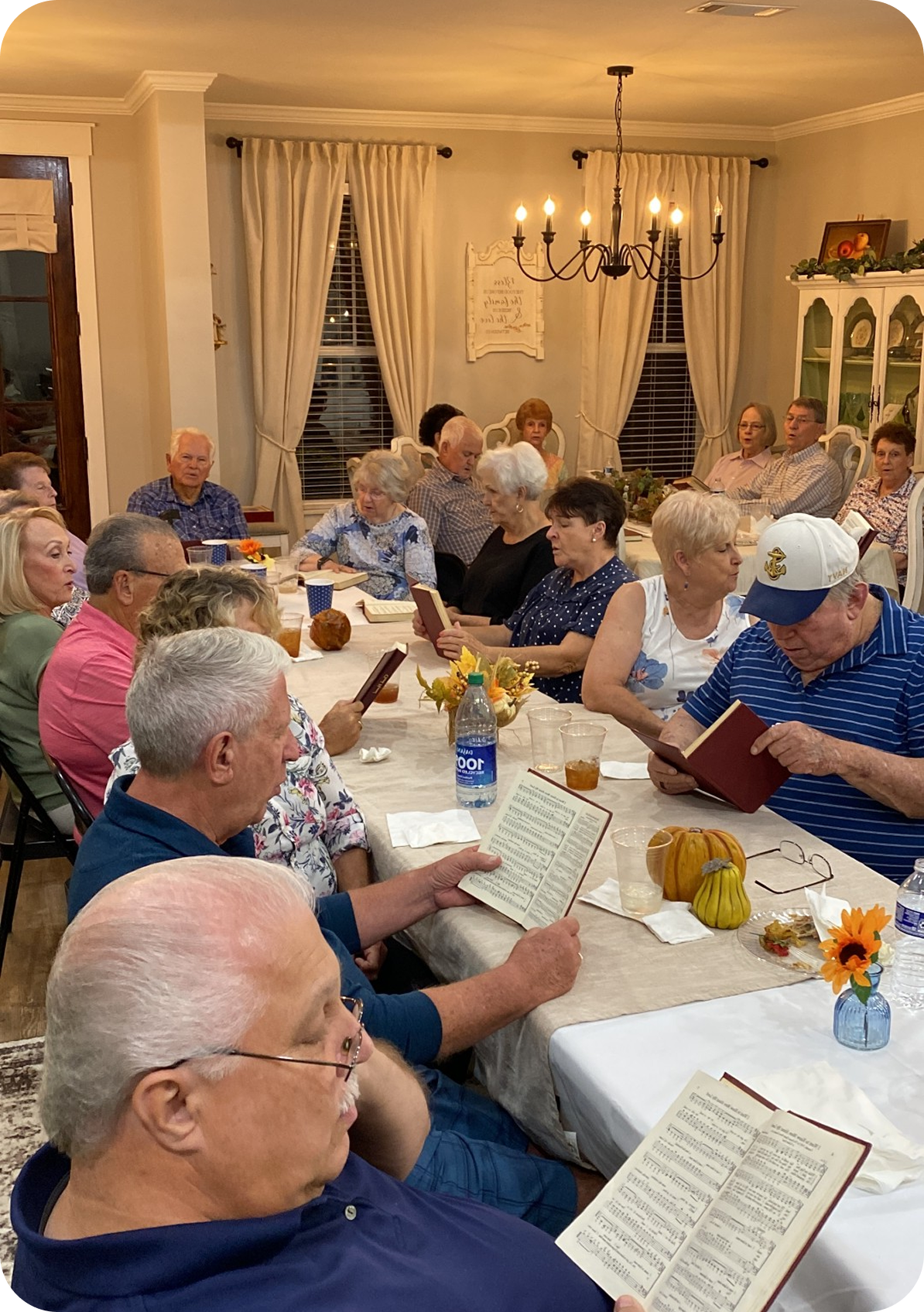 People seated at a long dining table in a warmly lit room, holding sheet music and singing.