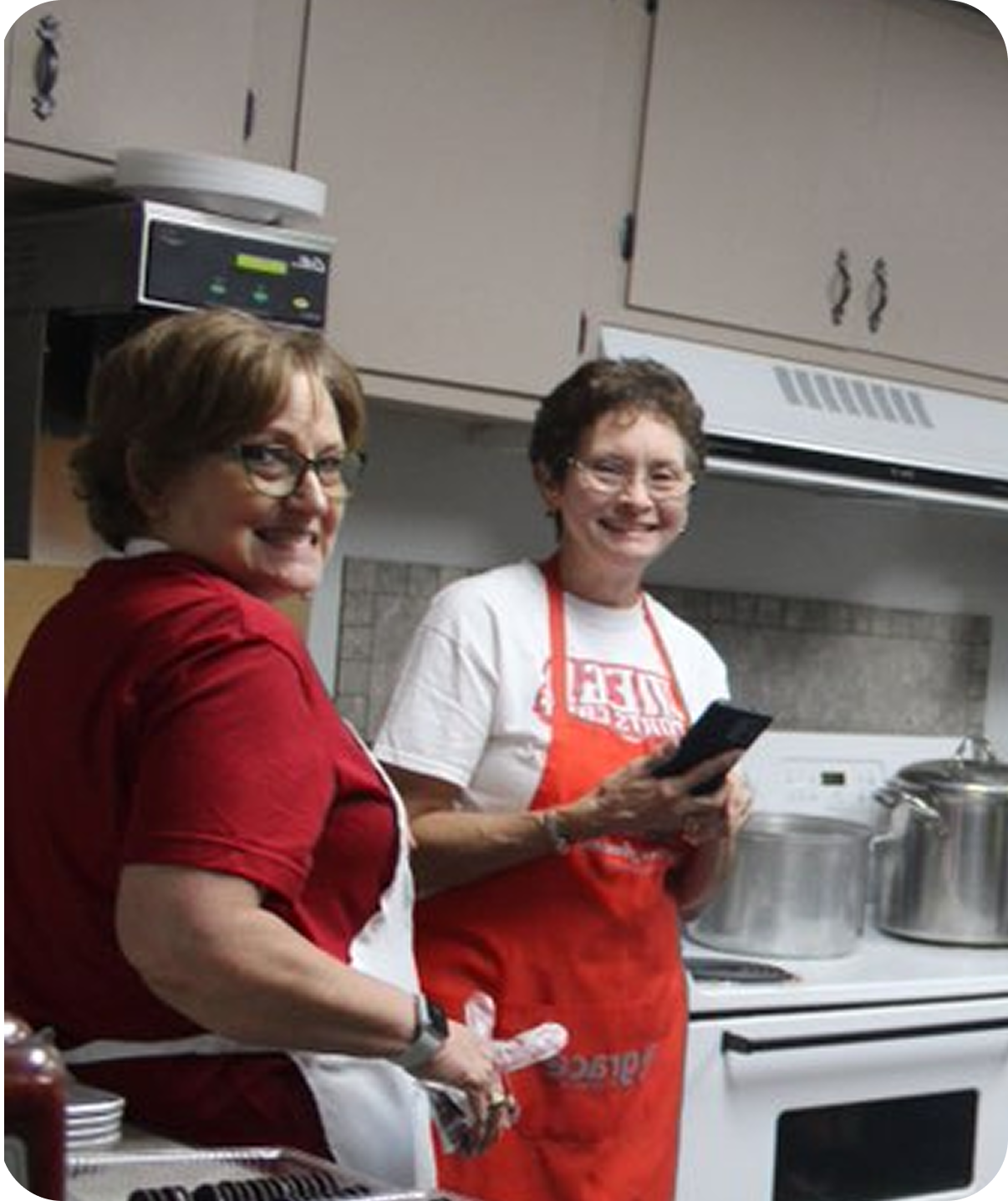 Two people in aprons smiling in a kitchen, one holding a black pan near a stove.