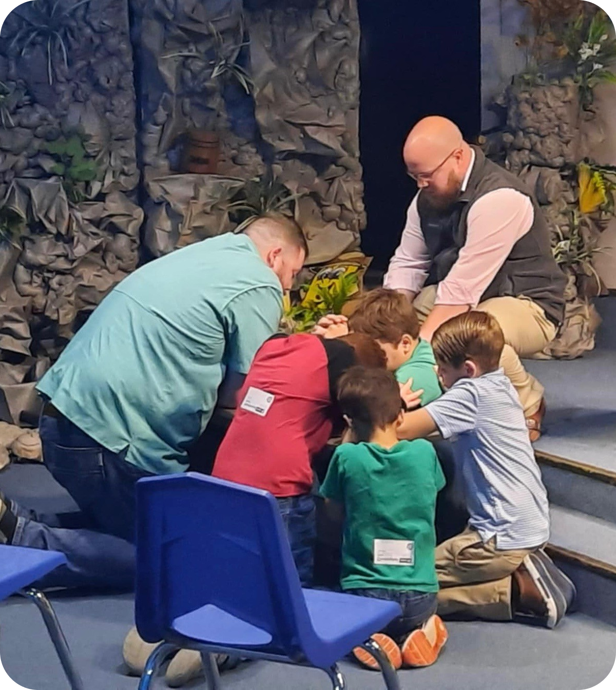 Teacher and children kneel around a small plant on a stage, with blue chairs nearby.