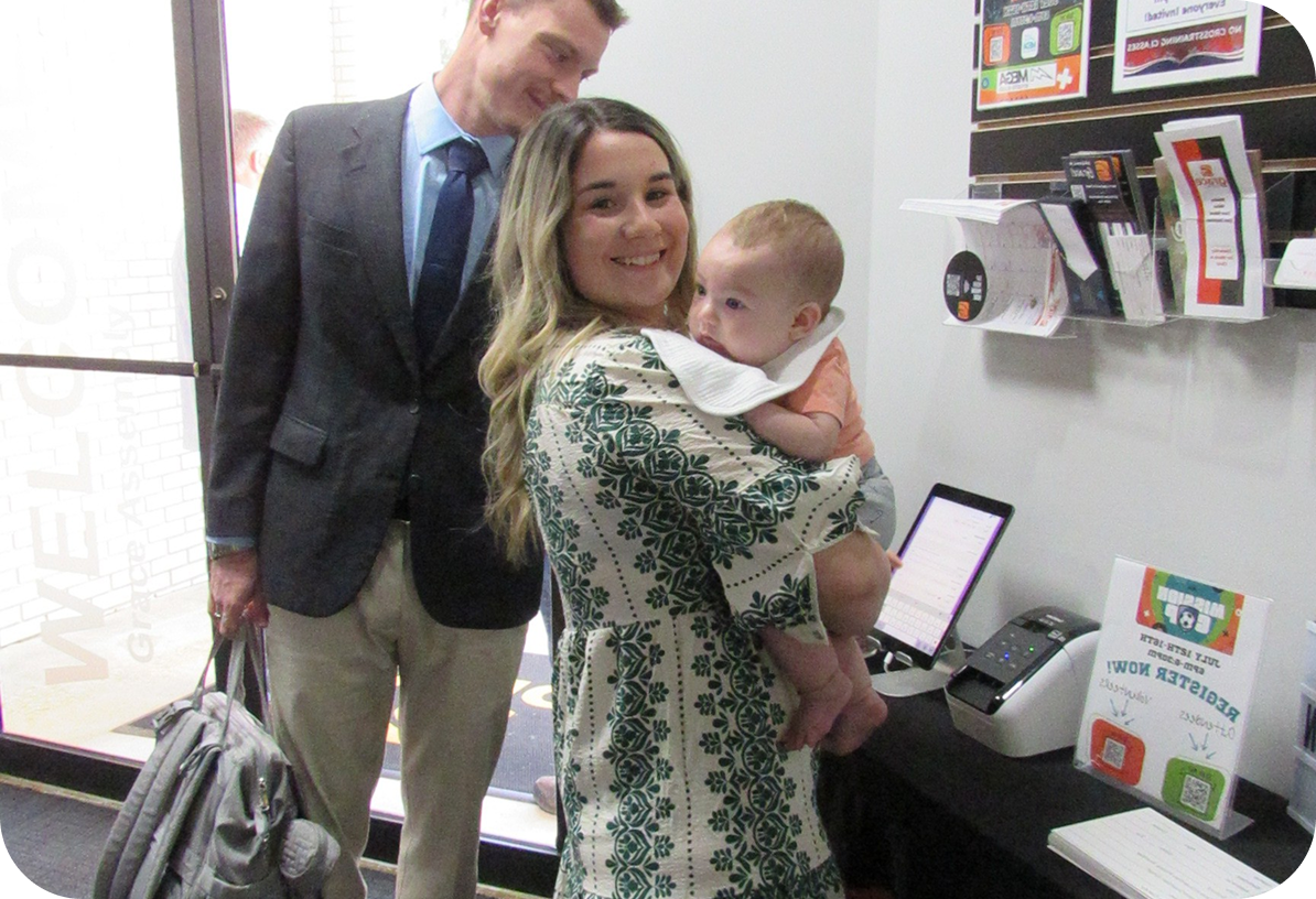 A couple holding a baby in a store or office, standing by a counter with brochures and a computer.