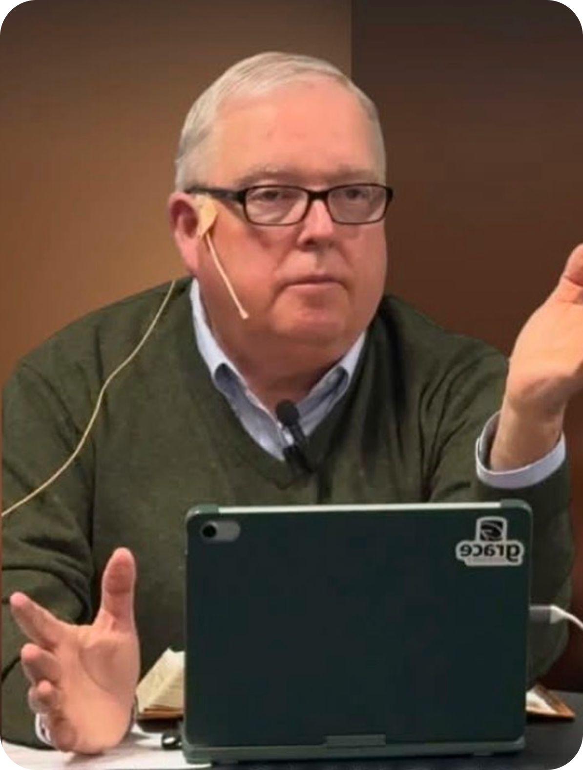 Man wearing glasses and green sweater speaking at a table with a tablet in front of him