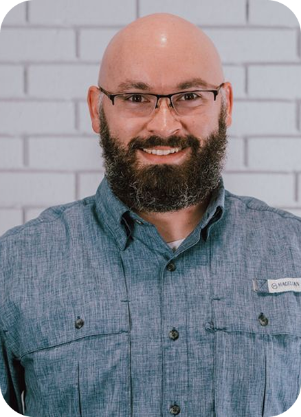 Smiling bald man with glasses and a beard in a blue button-up shirt against a white brick wall