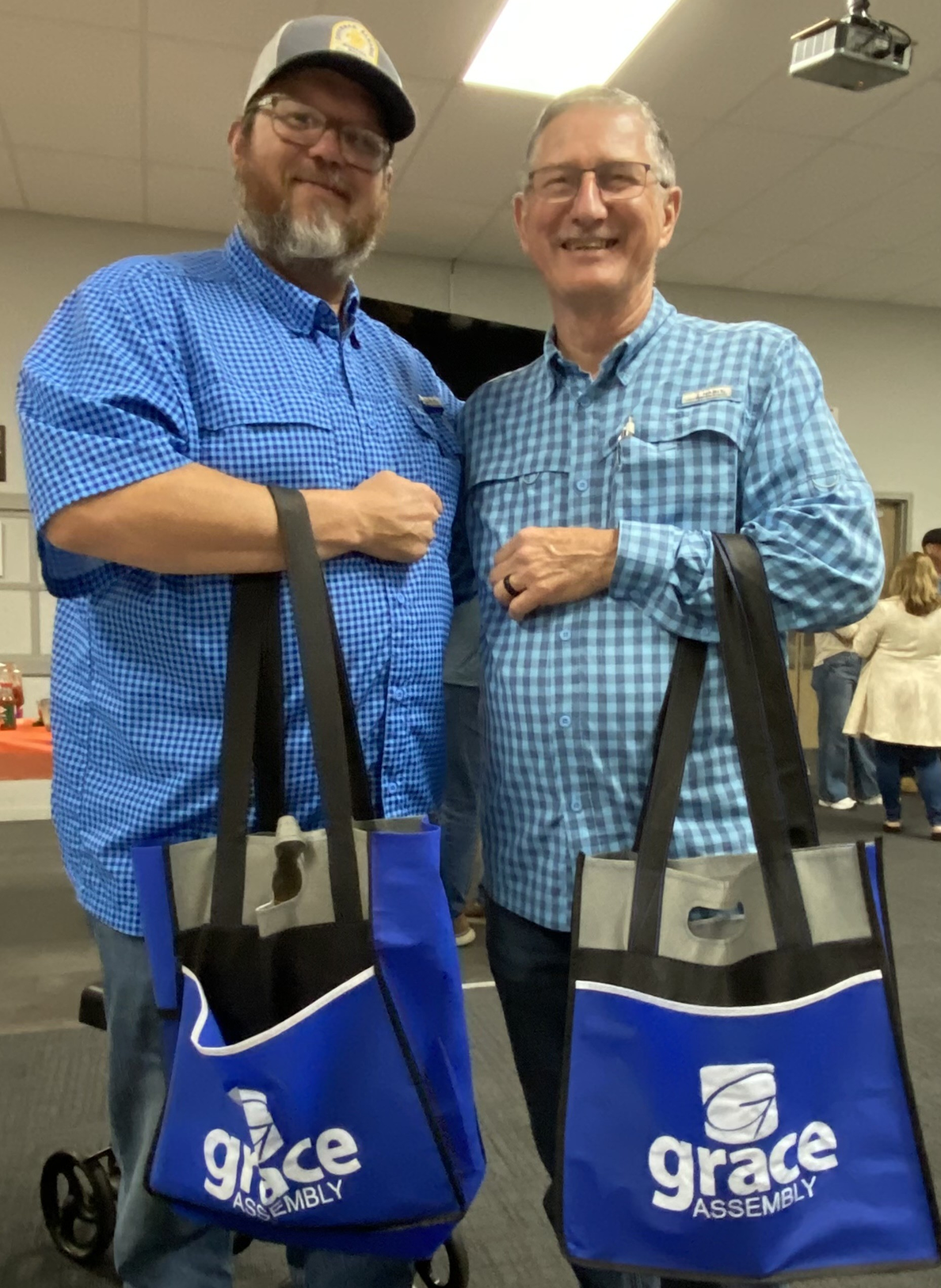 Two men smiling and holding blue Grace tote bags indoors