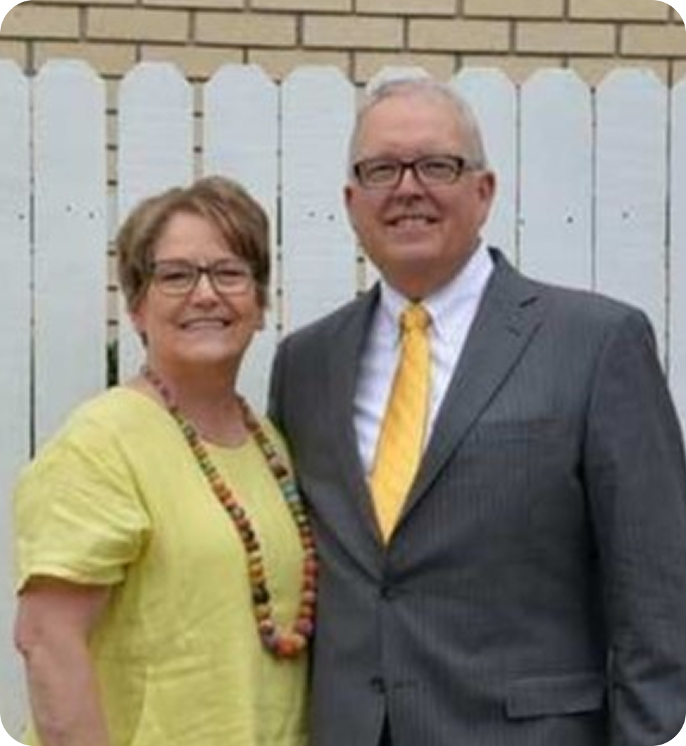 Smiling couple standing outdoors in front of a white fence, wearing glasses and formal attire.
