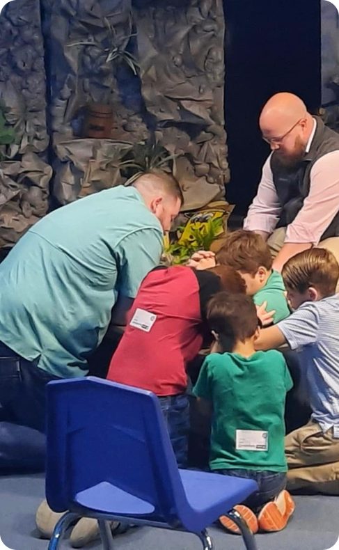 Adults and children gathered on the floor in a group activity, with a blue chair in the foreground.