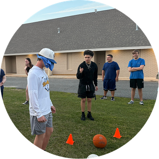 People gathered on a lawn around orange cones and a basketball in front of a building.