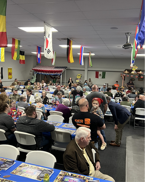 Crowded community hall with seated guests, hanging flags, and a stage decorated for an event.