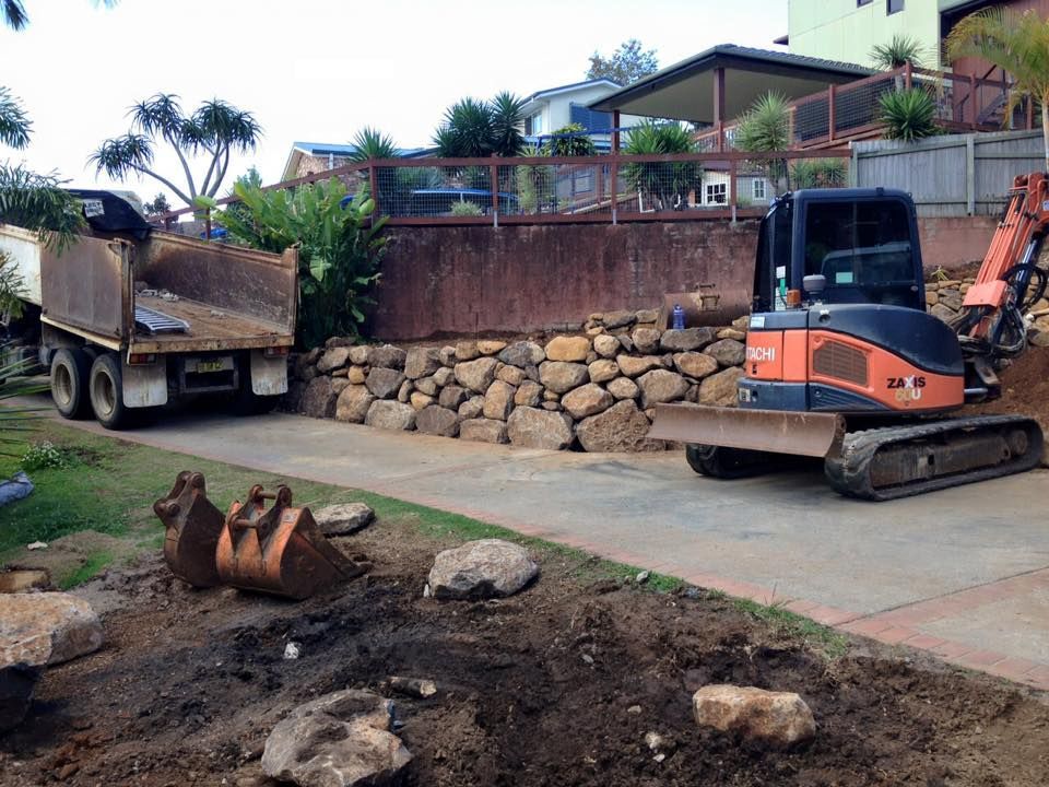 Excavator And Dump Truck At A Roadside Excavation Site With Exposed Dirt And A Stone Retaining Wall — Border Rockwalls in Stotts Creek, NSW