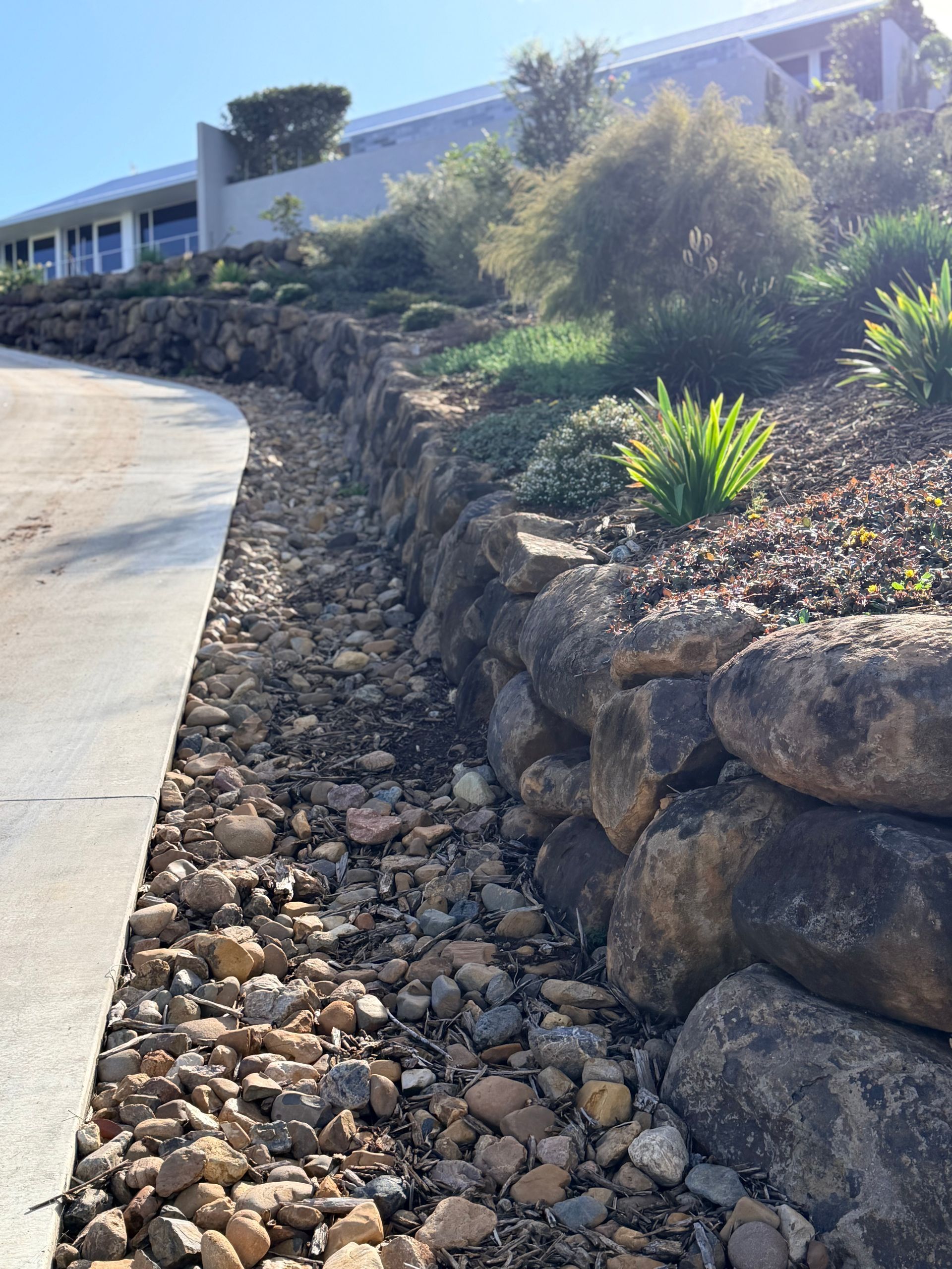 Rock-lined drainage ditch beside a curved driveway and landscaped hillside in bright sunlight —Border Rockwalls in Stotts Creek, NSW