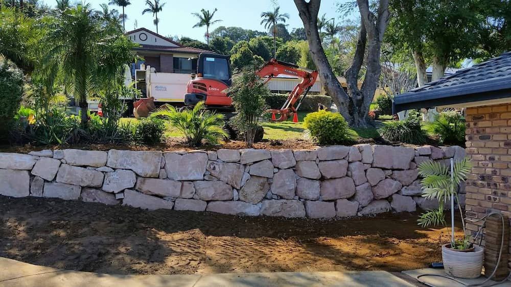 Stone Retaining Wall With An Orange Excavator And House On A Landscaped Hillside — Border Rockwalls in Gold Coast, NSW