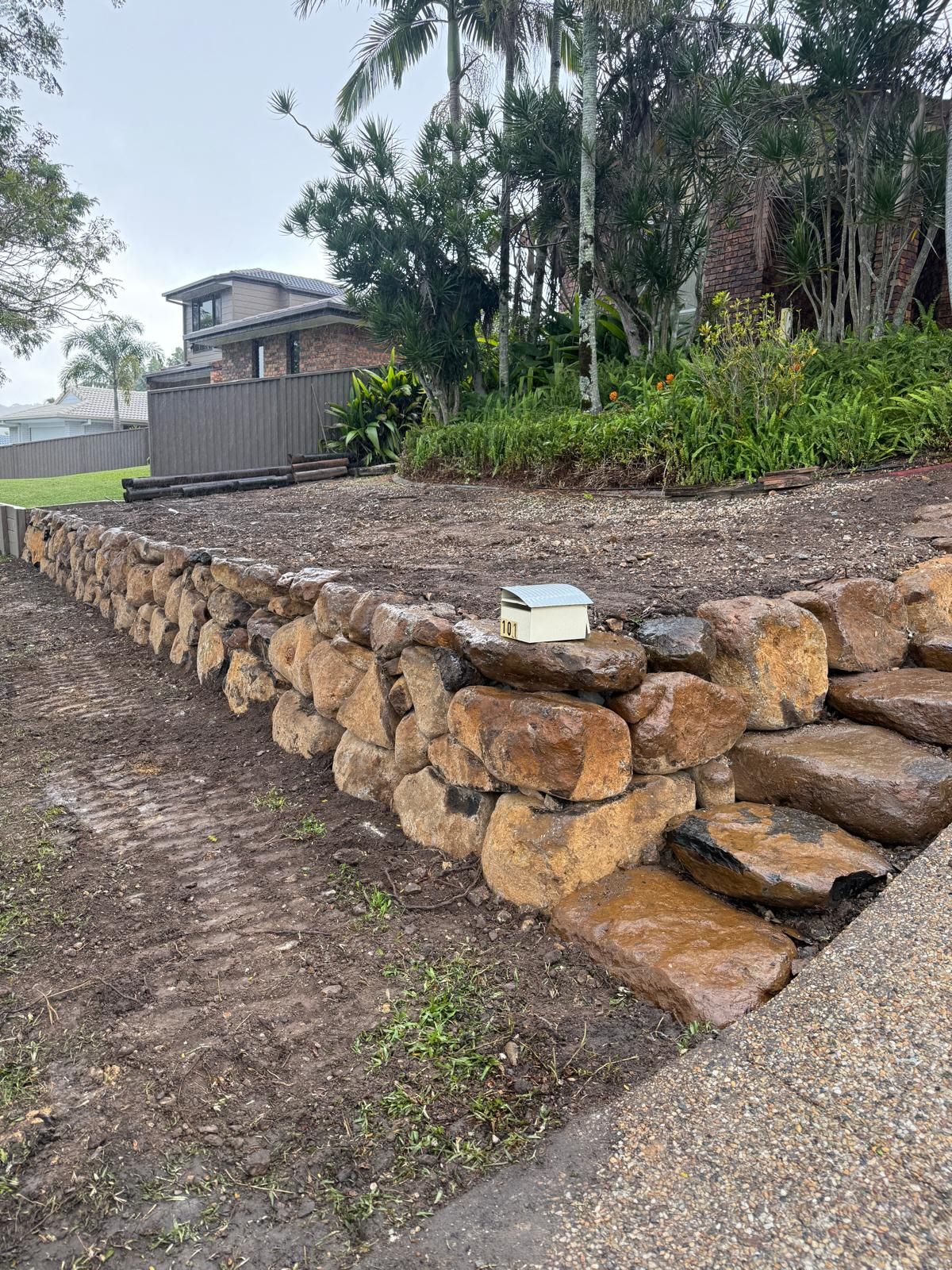 Stone retaining wall beside a muddy path, with shrubs and trees along a residential lot — Border Rockwalls in Stotts Creek, NSW