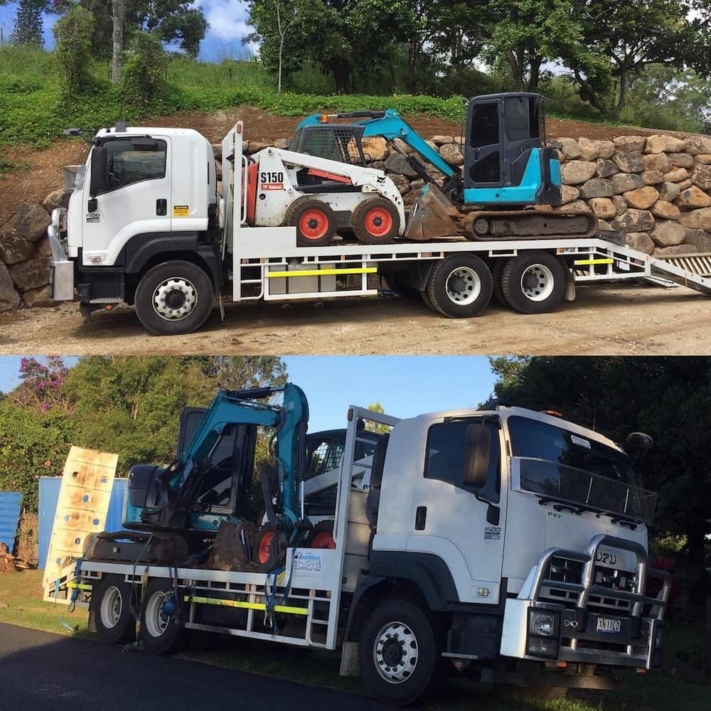 Two White Flatbed Trucks Loaded With Excavators At A Construction Site, One Outdoors And One By A Wall — Border Rockwalls in Stotts Creek, NSW