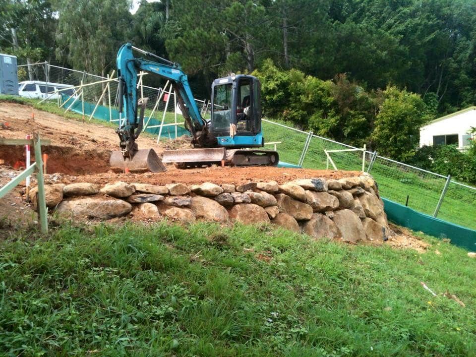 Excavator Working on a Stone Retaining Wall on a Grassy Hillside — Border Rockwalls in Stotts Creek, NSW