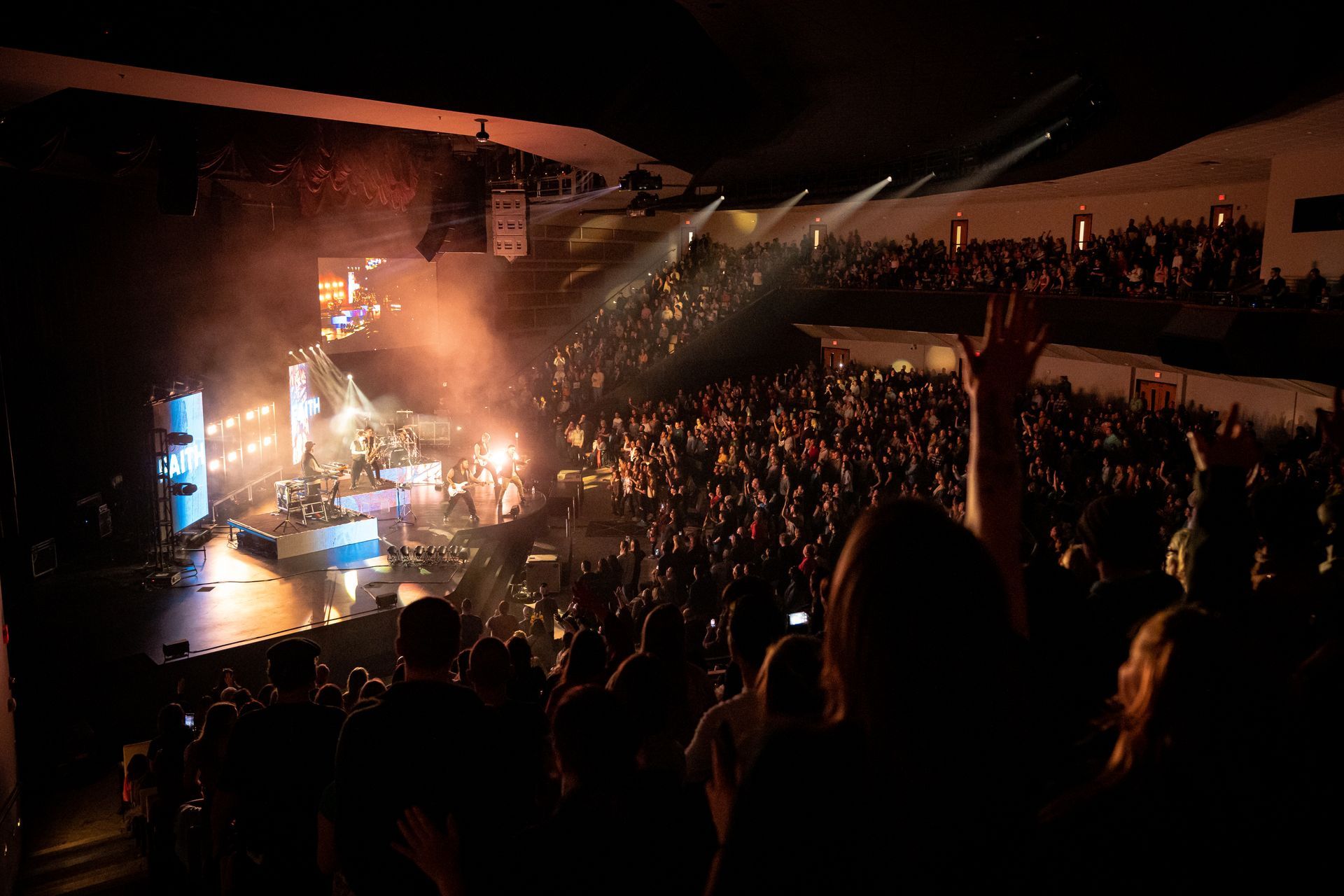 Concert crowd illuminated by stage lights. People cheer, one hand raised. Venue interior, dark.