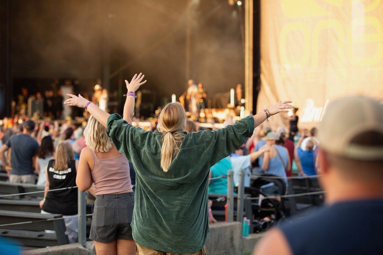 A crowd watches a concert outdoors. A person in a green sweater has arms raised in the air.