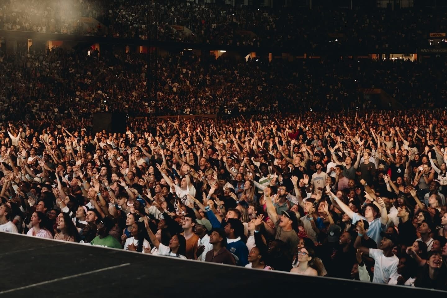 Large crowd at a concert raising hands in the air, lit by stage lights in a stadium.