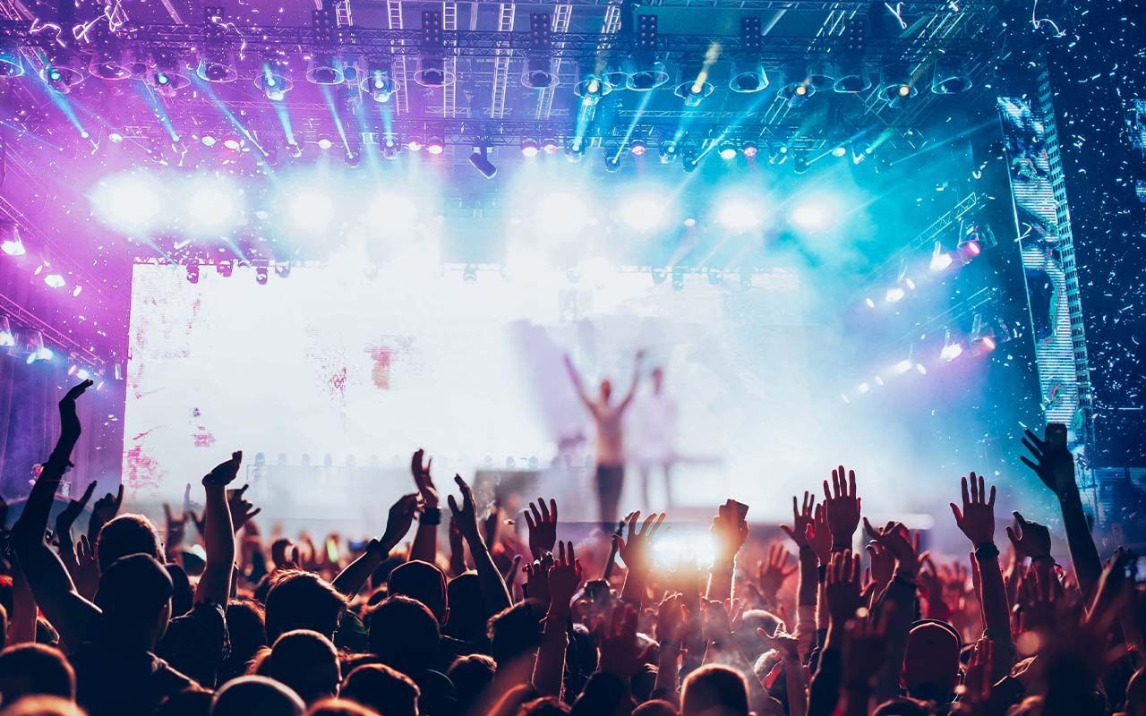 A crowd with raised hands enjoys a concert lit by bright purple and blue stage lights.