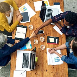 People around a wooden table shake hands during a meeting, laptops and papers are scattered about.