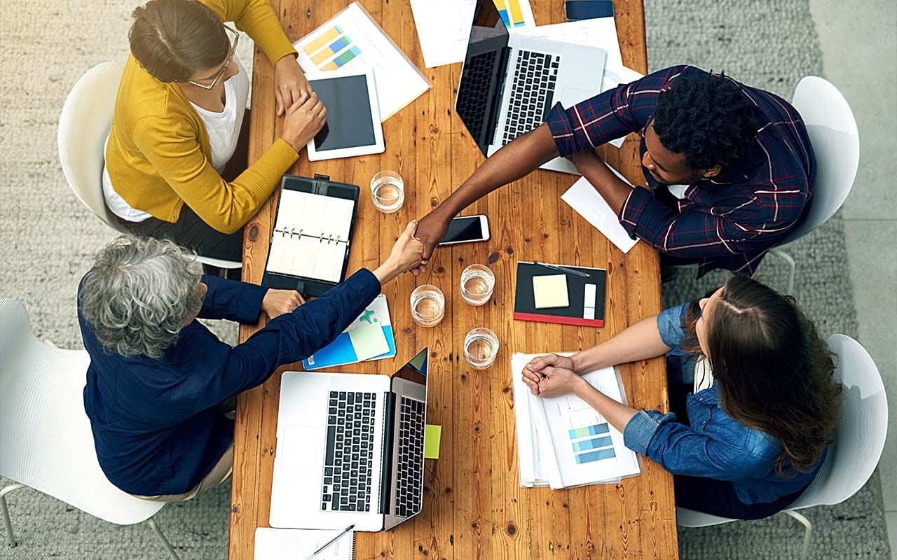 Four people shaking hands around a wooden table with laptops, papers, and glasses, in an office.