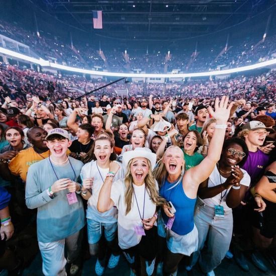 Large crowd at a concert; many are smiling and waving. Indoor arena lit up; stage not visible.