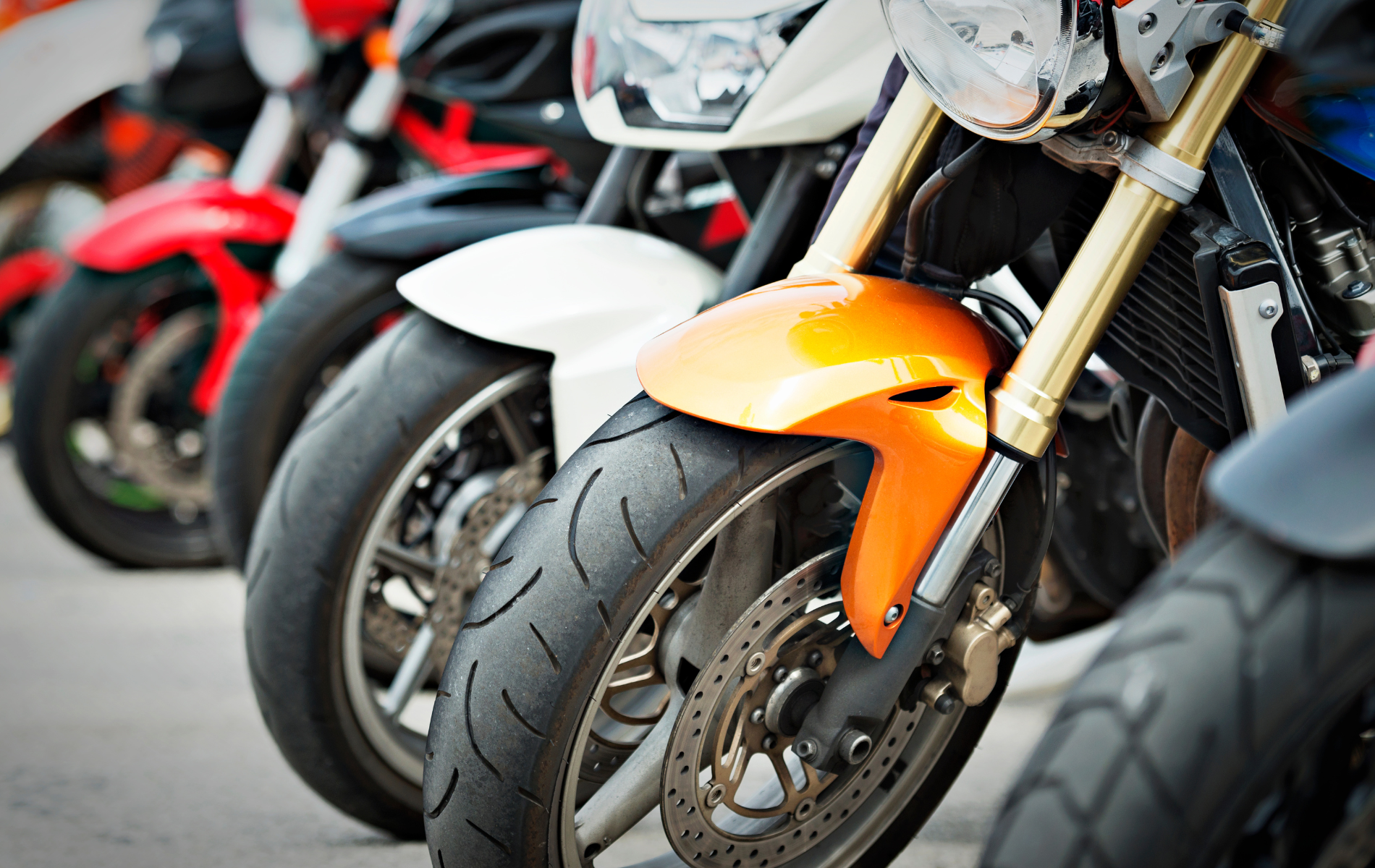 A row of parked motorcycles showing focus on their front tires, wheels, and suspension, with red, white, and orange fenders.