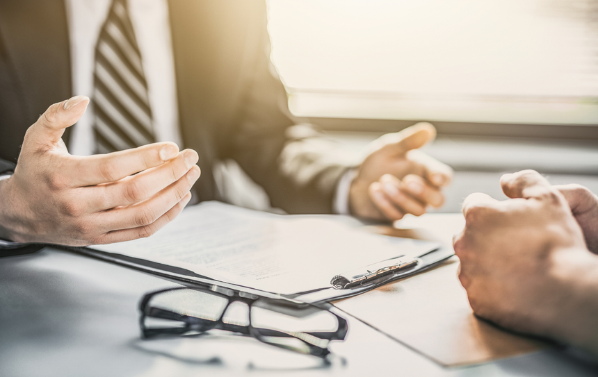 A person in a suit gesturing during a business meeting with a partner at a desk with papers and eyeglasses.