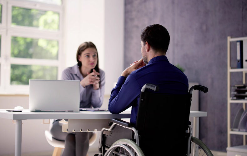 A person in a wheelchair sits across a desk from another individual in a professional office setting, facing each other.