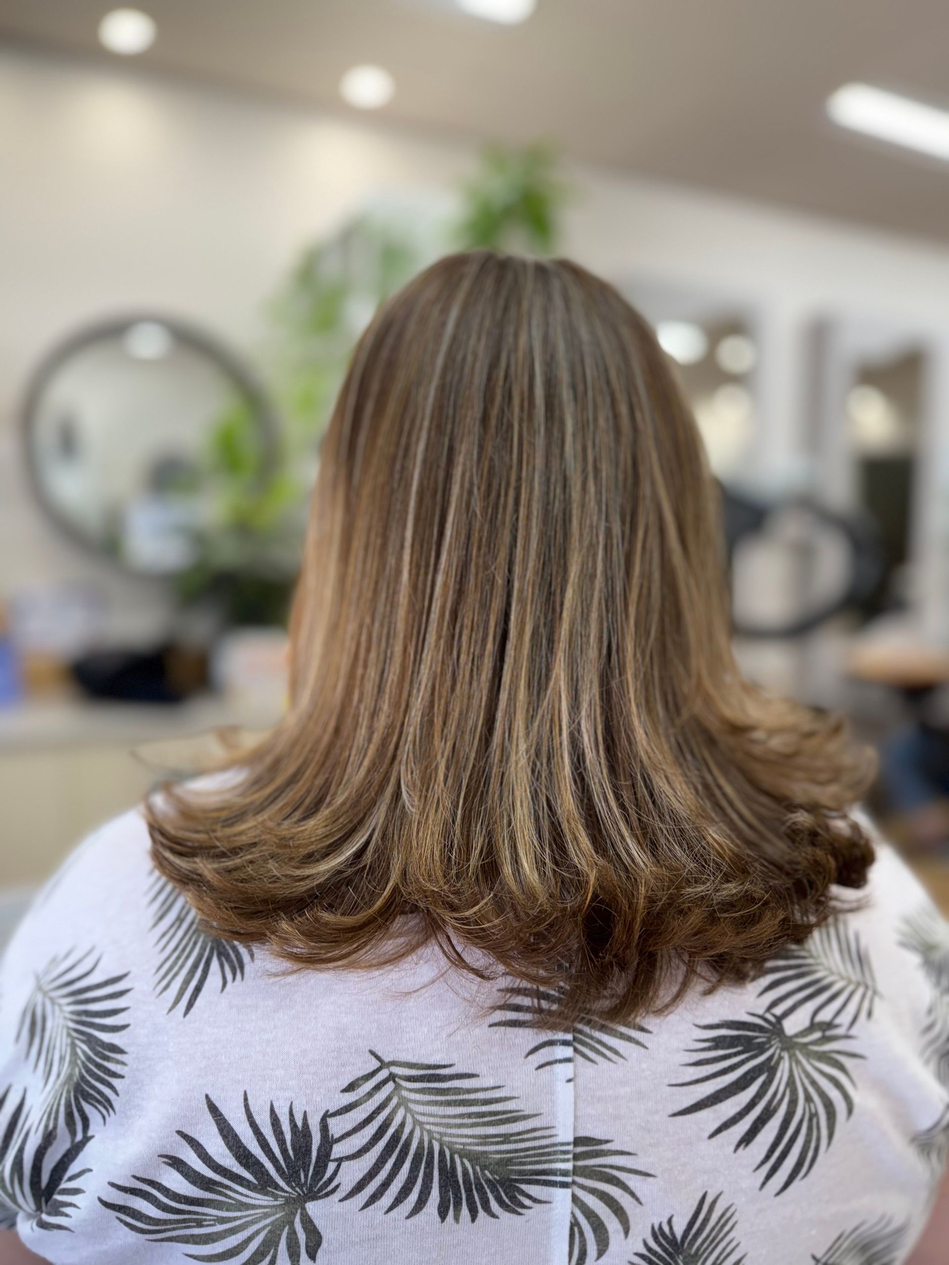 Back view of shoulder-length highlighted brown hair in a salon chair, with a leafy patterned shirt.