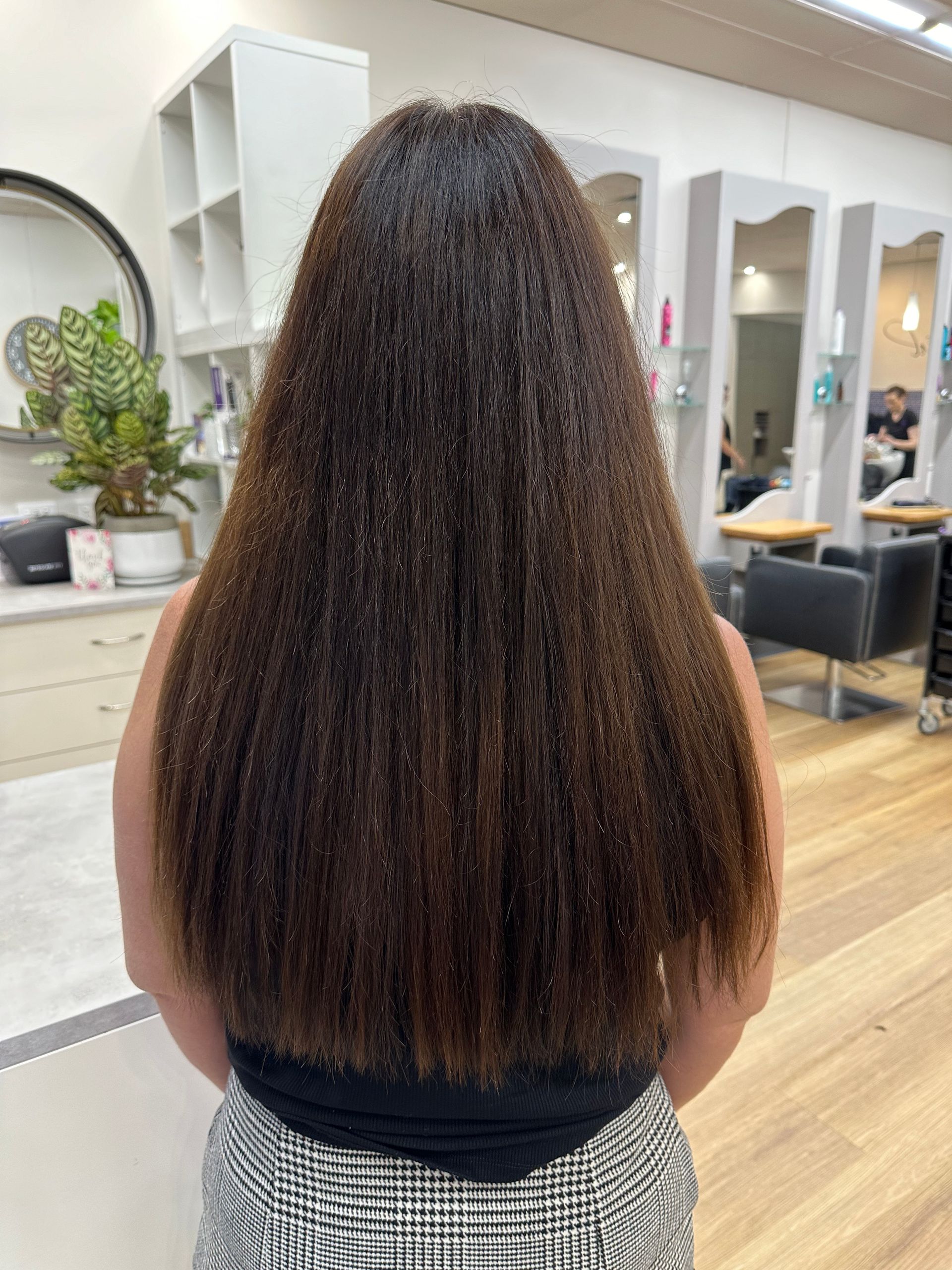 Back view of long, straight brown hair in a salon with mirrors and styling chairs
