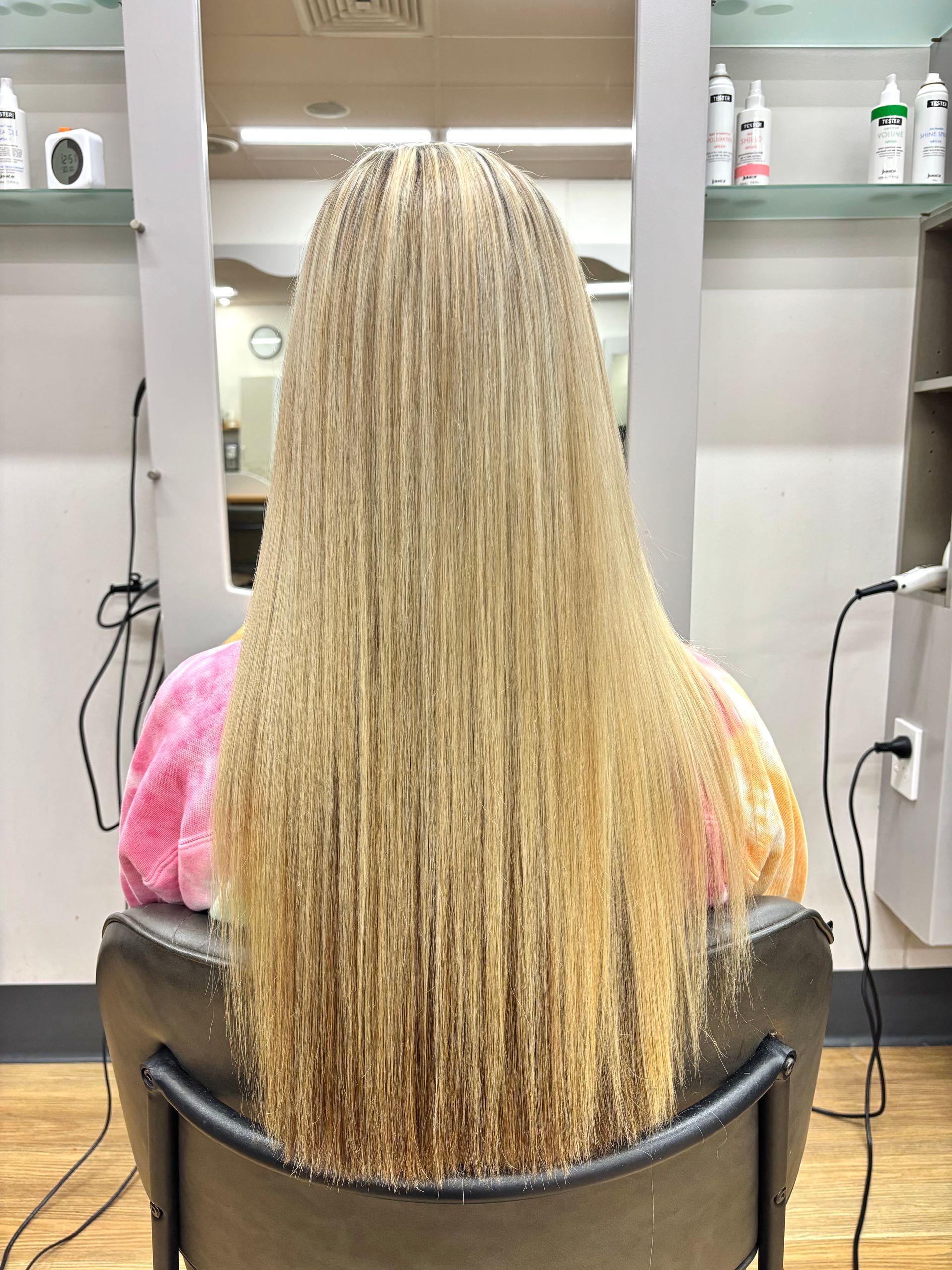 Person with long blonde braids seated in a salon chair, facing a mirror and wall shelves.