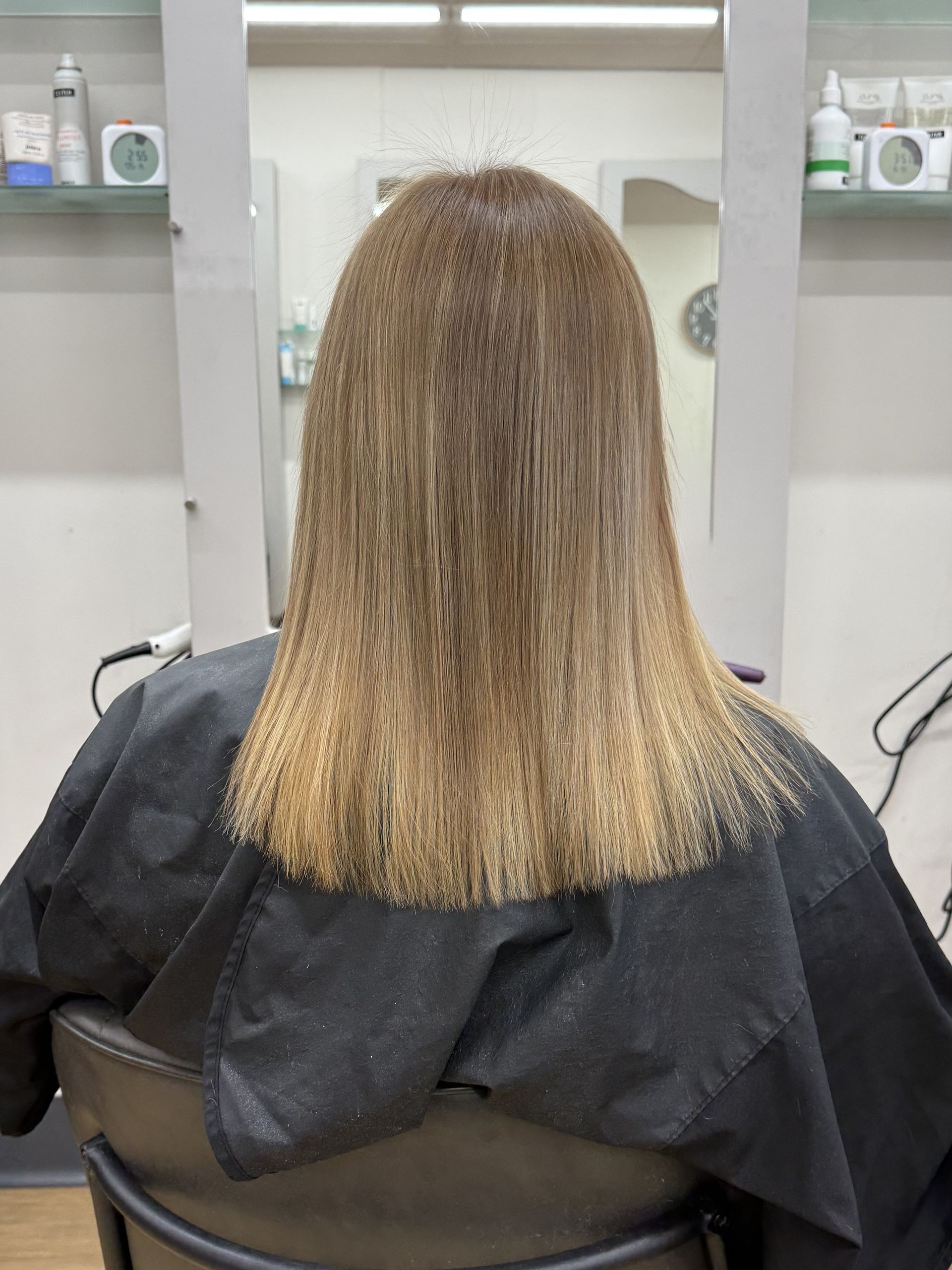 Person with blonde hair seated in a salon chair, viewed from behind, with mirrors and shelves in the background.