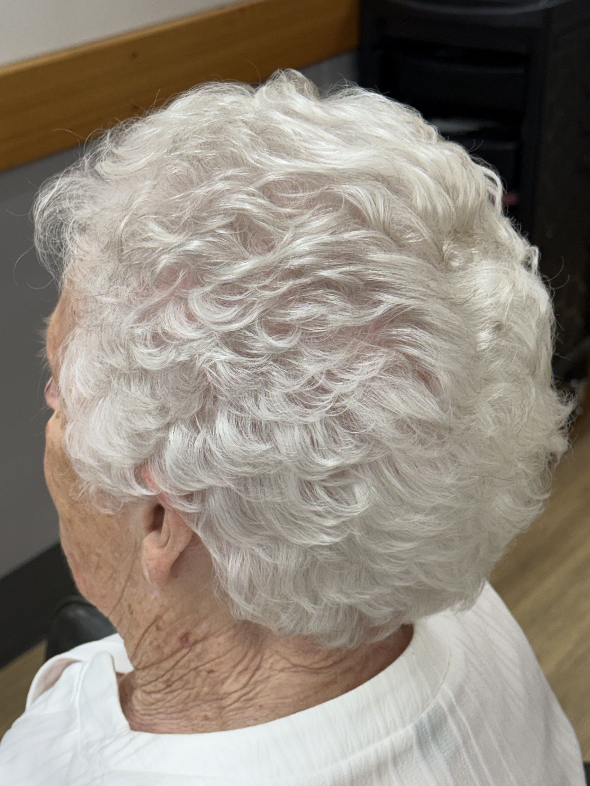 Close-up of a person with short curly white hair, seen from behind in a white shirt.