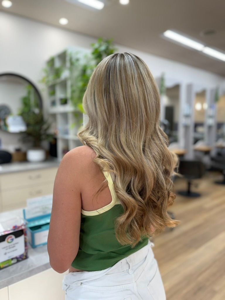 Woman with long blonde hair standing at a salon counter, facing the bright, modern interior.