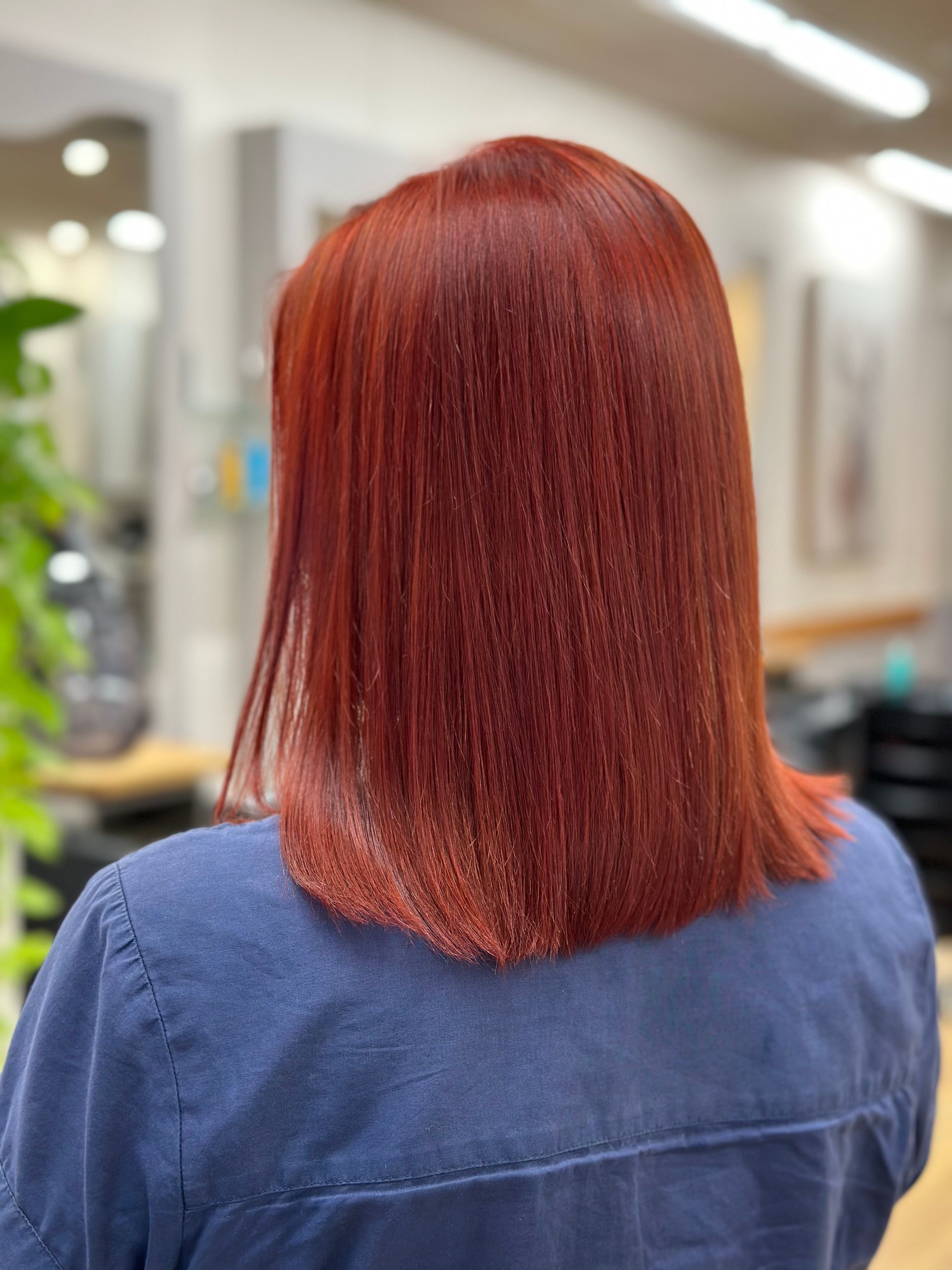 Back view of a person with straight red hair in a blue shirt, in a salon setting