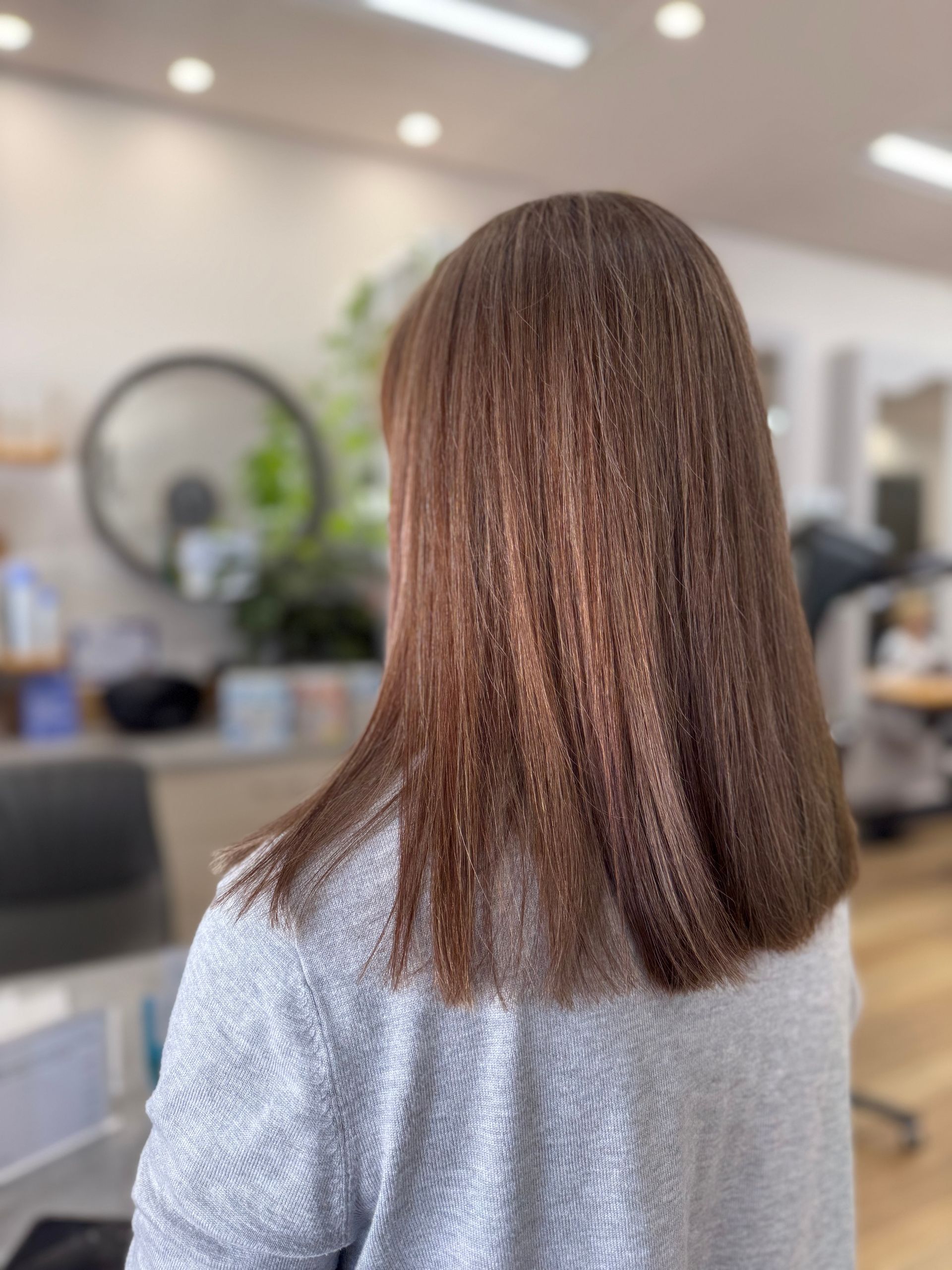 Woman with straight brown hair in a salon chair, shown from behind with softly lit salon background.