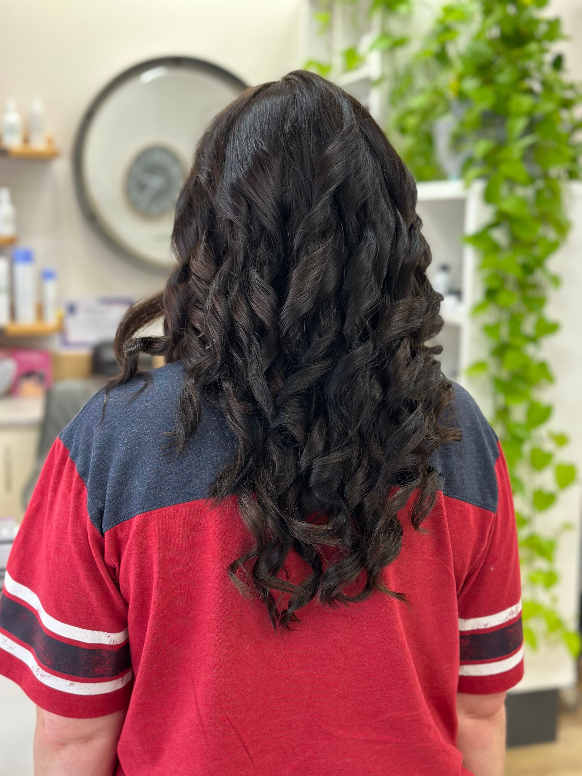 Woman with long curly hair seen from behind in a red and navy top, standing indoors near a mirror and plant.