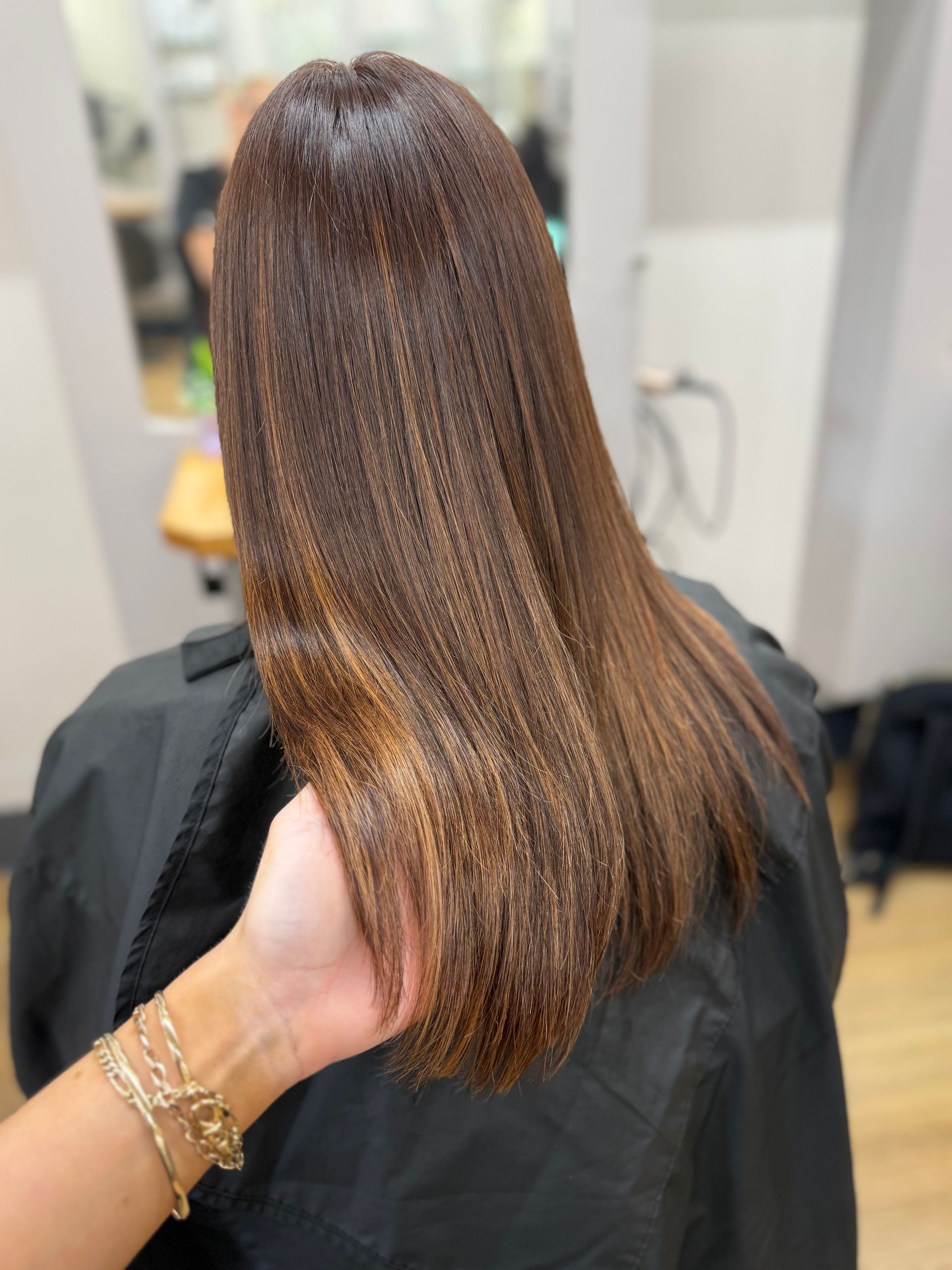 Back view of straight brown hair with subtle highlights, held by a hand in a salon chair
