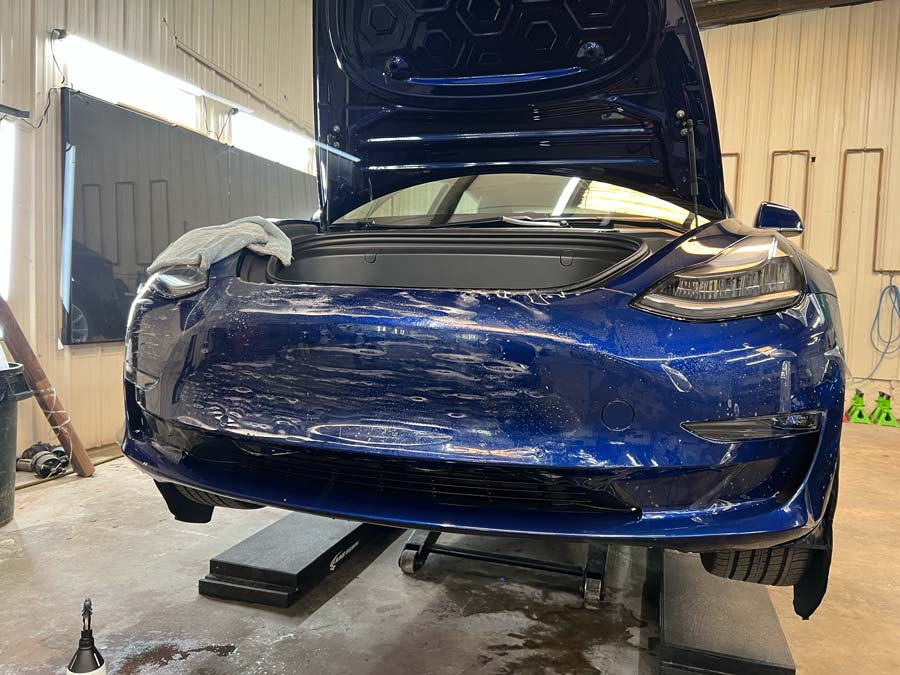 A blue tesla model 3 with the hood open is sitting on a lift in a garage.
