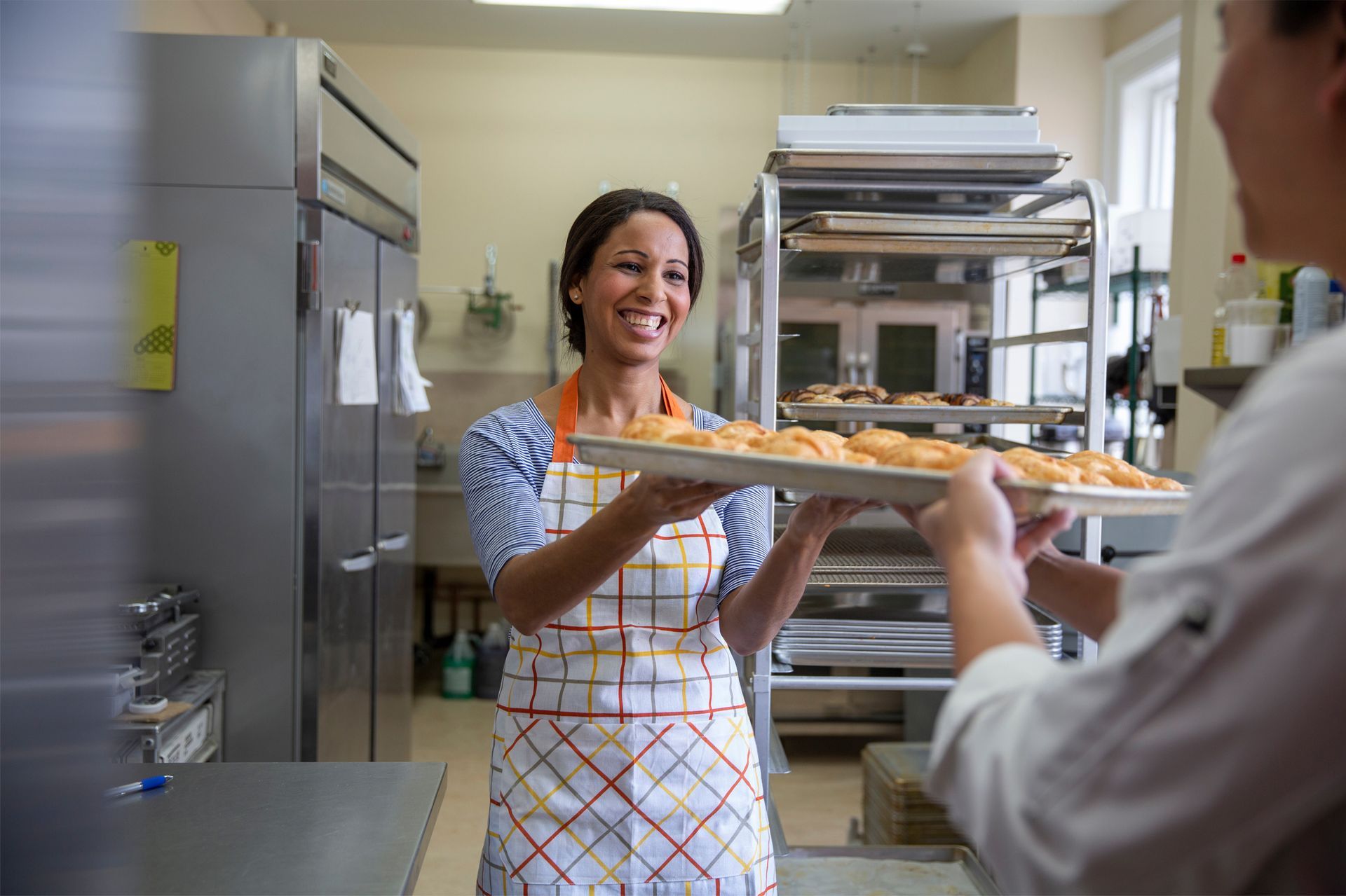 Woman in a kitchen handing a tray of food to someone, smiling while wearing an apron