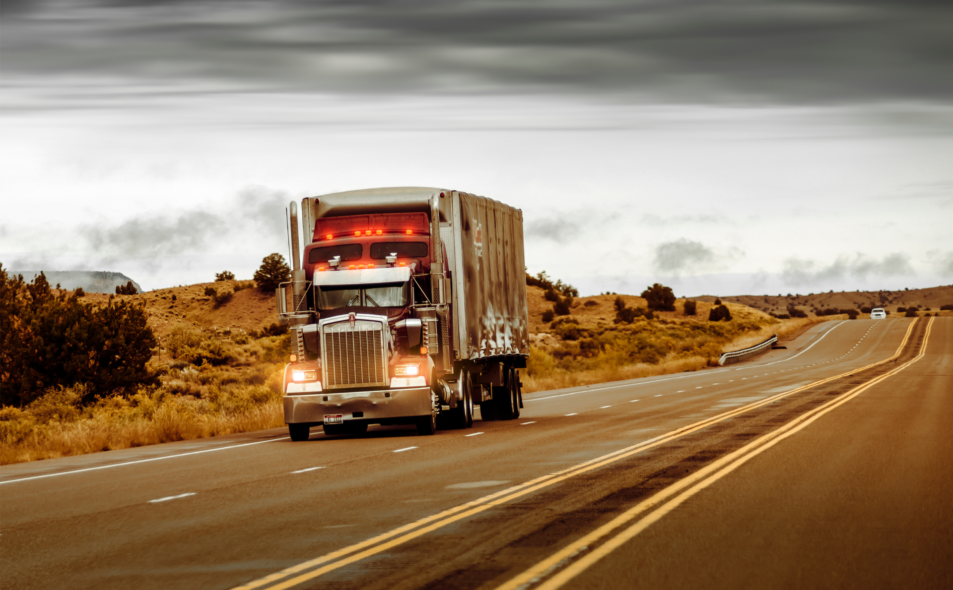 Semi-truck driving on a deserted highway through a dry, open landscape under a cloudy sky
