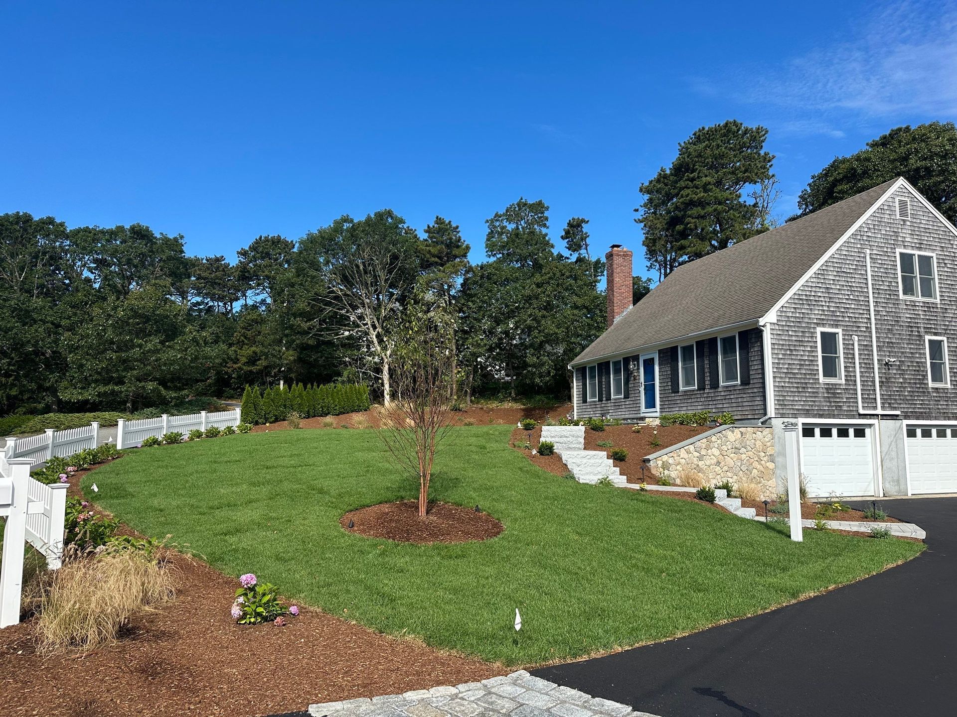 A shingled house with a white garage sits next to a green lawn, a young tree, and a white fence under a clear blue sky.