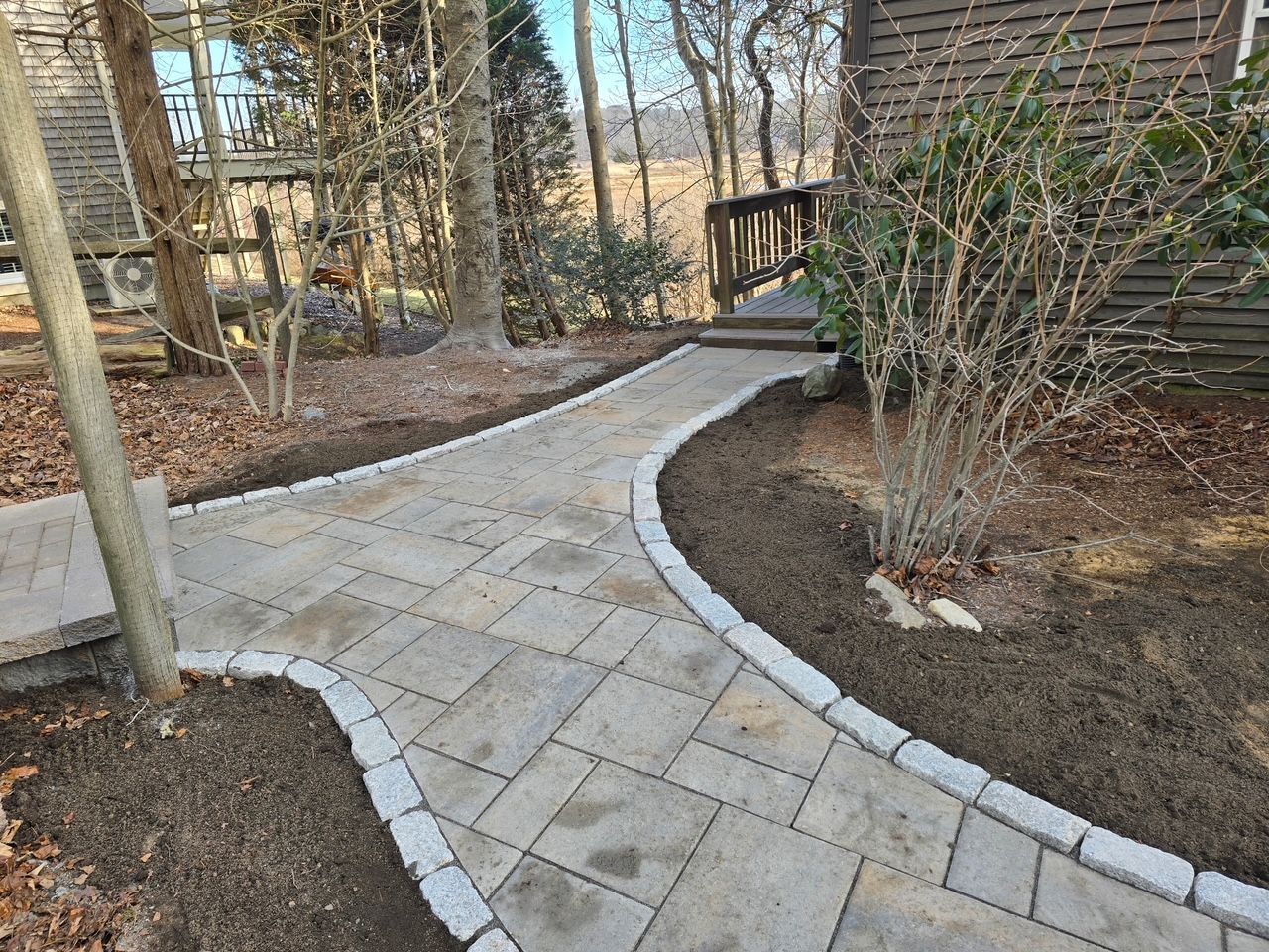A stone paver walkway with a gray border winds through a yard towards wooden stairs leading to a house entrance.